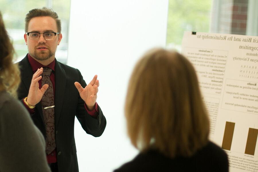 Male student in front of research poster gesturing and talking to two individuals.