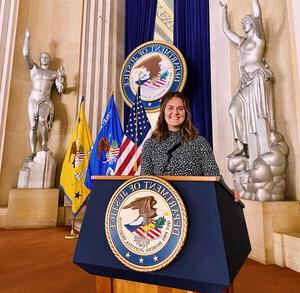 一名年轻女子站在讲台后面，讲台上有一个大印章，上面写着“司法部”，她身后是一间大房间的雕像.