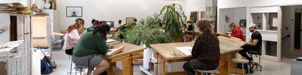 Art students drawing and sitting at large wooden drawing desks in circle around plants.